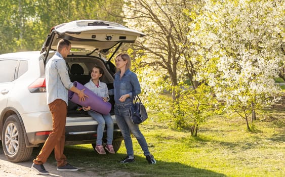 Happy family standing together near a car with open trunk enjoying view of rural landscape nature. Parents and their kid leaning on vehicle luggage compartment. Weekend travel and holidays concept