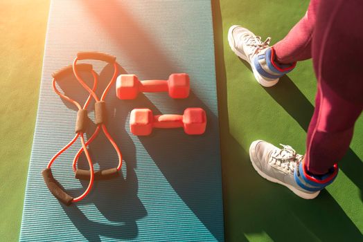 Woman legs near mat with expander and two dumbbells