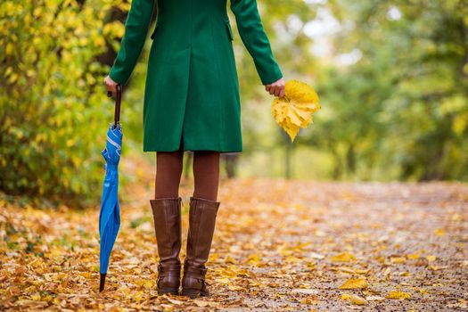 Woman in boots holding umbrella and fall leaves while standing in the park.