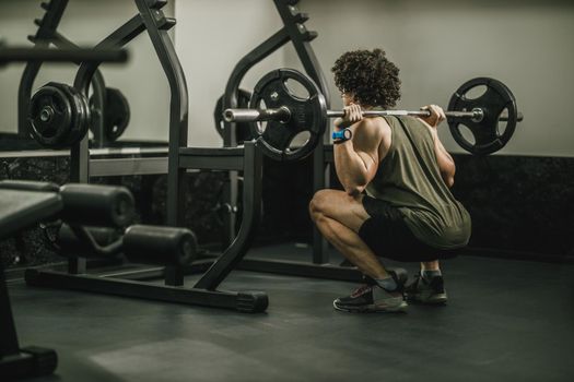 Shot of a muscular guy in sportswear working out at the hard training in a gym. He is doing squat exercises with barbell.