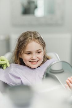 A cute little girl looking her teeth in a mirror after dental procedure at dental clinic.