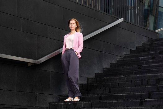 Young serious beautiful blonde millennial woman with long hair in pink elegant clothes stands on steps near modern building. Selective focus.