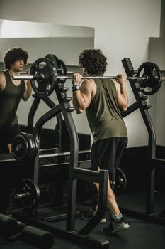 Shot of a muscular guy in sportswear working out at the hard training in a gym. He is doing squat exercises with barbell.