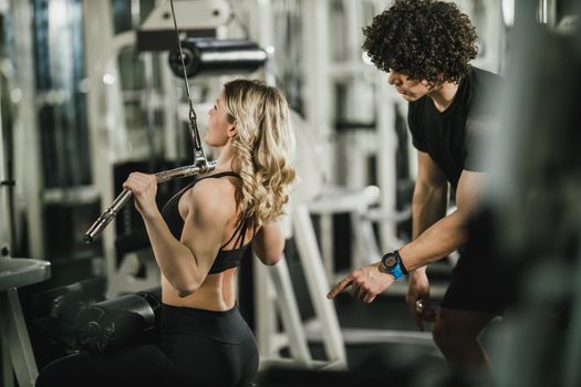 A young muscular woman is doing training at machine supporting by coach in the gym.