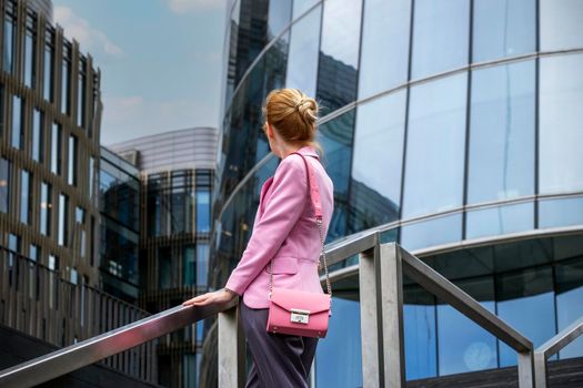 Young blonde woman with strict hairstyle in pink jacket stands on steps with her back to camera and looks at modern office building made of glass and steel. Selective focus.