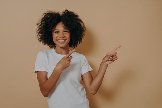 Positive cheerful dark skinned woman dressed in white basic tshirt pointing at copy space isolated on beige background, advertising displaying product, showing place for your advertisement promotion