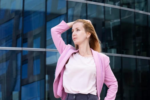 Young beautiful blonde woman with long hair in pink elegant clothes stands near a modern building made of glass and steel. Selective focus.