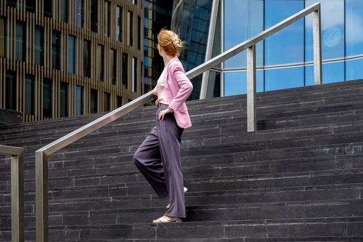 Young blonde of millennium with strict hairstyle in pink elegant clothes stands on steps near modern office building in full growth, half-turned to camera. Selective focus.