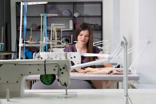 Young dressmaker woman sews clothes on sewing machine. Smiling seamstress and her hand close up in workshop. Focus on sewing machine and tissue.