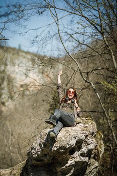 A cheerful young woman sitting on the top of the mountain cliff and having fun the outdoors.