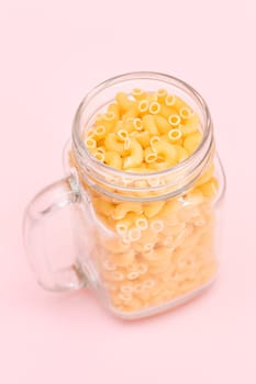 Uncooked Chifferi Rigati Pasta in Glass Jar on Pink Background. Fat and Unhealthy Food. Scattered Classic Dry Macaroni. Italian Culture and Cuisine. Raw Pasta