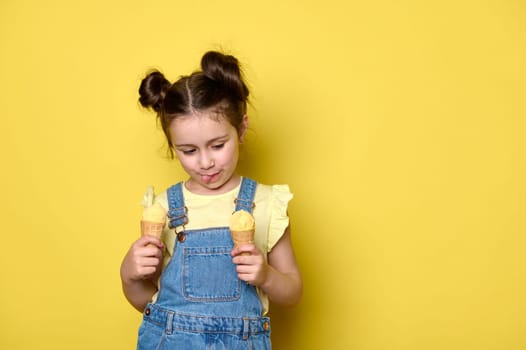 Beautiful Caucasian baby girl with trendy hairstyle, showing tongue while licking delicious ice cream in waffle cone, dressed in casual blue denim and yellow t-shirt, isolated on yellow background.