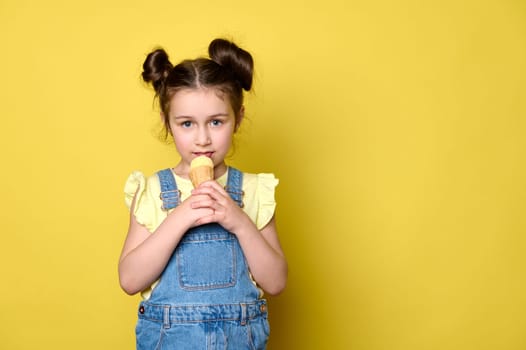 Lovely baby girl tasting delicious ice cream scoop in waffle cone, looking at camera, isolated over yellow background with free ad space. Summer holidays. Happy childhood. Fashionable kids concept