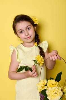Vertical studio portrait of beautiful little child girl holding a purple wicker basket with yellow rose flowers, smiling looking at camera, isolated over yellow color background. Happy Mother's Day