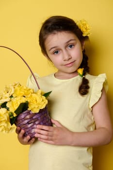 Lovely little girl with flowers in pigtail, dressed in yellow clothes, holding purple wicker basket with yellow roses, smiling a beautiful toothy smile, looking at camera, isolated yellow background