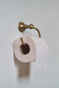 Pink toilet paper hanging on a gold toilet paper holder on a light bathroom wall. High quality photo