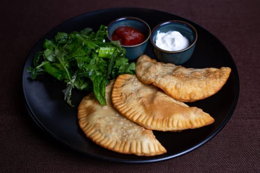 fried chebureks with sauce and herbs on a black plate.