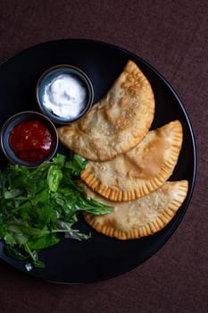 top view fried chebureks with sauce and herbs on a black plate.