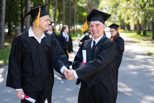 A group of graduates in robes outdoors. An elderly man and a young guy congratulate each other on receiving a diploma
