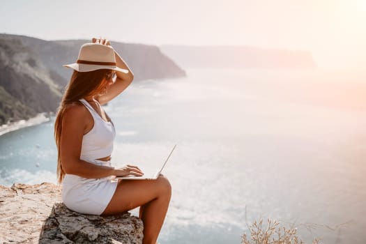 Successful business woman in yellow hat working on laptop by the sea. Pretty lady typing on computer at summer day outdoors. Freelance, travel and holidays concept.