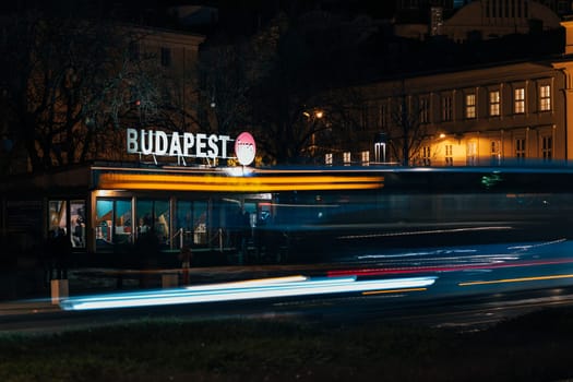 Reflections of illuminated buildings create a stunning backdrop for a bustling city evening. A bus and its passengers pause at a stop, amidst grand architecture in Budapest.