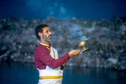 Rishikesh, India - 29th Apr'23: : Priest in red and white praying to the holy river ganga with a lit oil diya lamp giving off smoke as part of the evening aarti prayer performance at triveni ghat