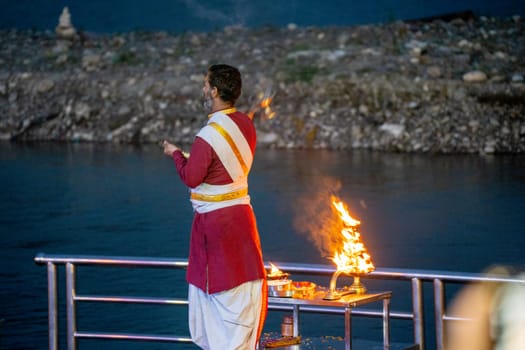 Rishikesh, Uttarakhand - circa 2023 : Priest in red and white praying to the holy river ganga with a lit oil diya lamp giving off smoke as part of the evening aarti prayer performance at triveni ghat