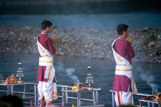 Priest in red and white chanting hymns to the holy river ganga with a lit oil diya lamp giving off smoke as part of the evening aarti prayer performance at triveni ghat