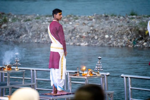 Rishikesh, Uttarakhand - circa 2023 : Priest in red and white chanting hymns to the holy river ganga with a lit oil diya lamp giving off smoke as part of the evening aarti prayer performance at triveni ghat