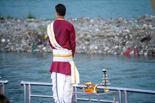 Priest in red and white chanting hymns to the holy river ganga with a lit oil diya lamp giving off smoke as part of the evening aarti prayer performance at triveni ghat