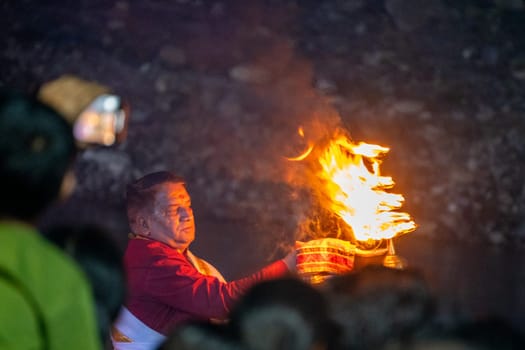Rishikesh, India - 29th Apr'23: Priest in red and white praying to the holy river ganga with a lit oil diya lamp giving off smoke as part of the evening aarti prayer performance at triveni ghat India