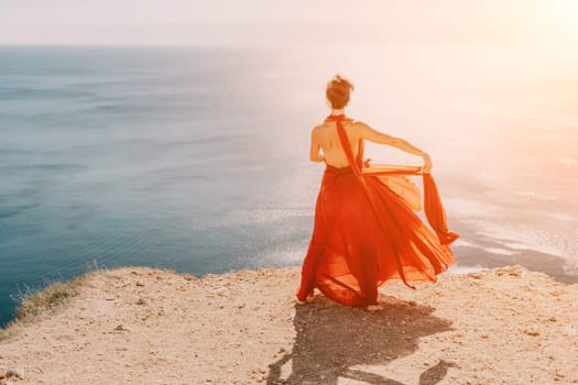 Side view a Young beautiful sensual woman in a red long dress posing on a rock high above the sea during sunrise. Girl on the nature on blue sky background. Fashion photo.