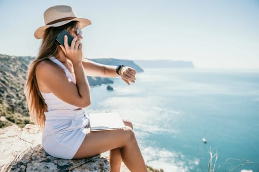 Successful business woman in yellow hat working on laptop by the sea. Pretty lady typing on computer at summer day outdoors. Freelance, travel and holidays concept.