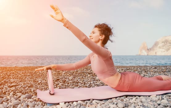 Middle aged well looking woman with black hair doing Pilates with the ring on the yoga mat near the sea on the pebble beach. Female fitness yoga concept. Healthy lifestyle, harmony and meditation.