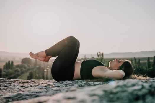 Well looking middle aged woman with long hair, fitness instructor in leggings and tops doing stretching and pilates on the rock near forest. Female fitness yoga routine concept. Healthy lifestyle.