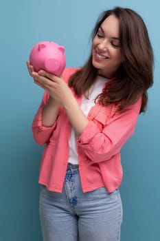 positive brunette young woman in a shirt and jeans puts her budget into a piggy bank.