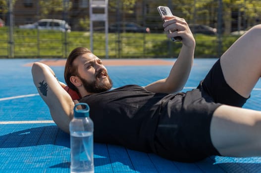 Portrait of tired athletic lebanese man in sportswear after playing basketball game. Guy lying down on ball relaxing use smartphone texting share messages on urban city court. Fitness on sports field