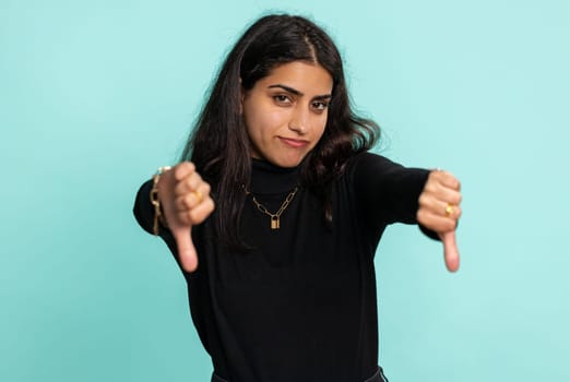 Dislike. Upset unhappy indian woman in sweater showing thumbs down sign gesture, expressing discontent, disapproval, dissatisfied, dislike. Pretty young girl. Indoor studio shot on blue background