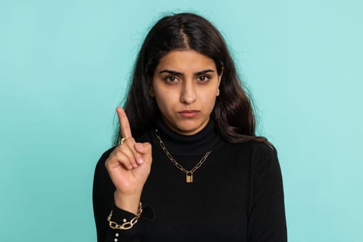 Indian pretty woman shakes finger and saying No, be careful, scolding, giving advice to avoid danger mistake, disapproval sign. Young girl isolated alone on blue studio background. People lifestyle