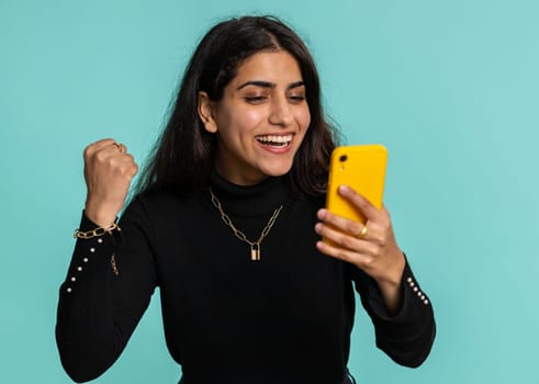 Happy excited joyful indian woman in black sweater use smartphone typing browsing shouting say wow yes found out great big win, good news, lottery goal achievemen, celebrating success, winning game