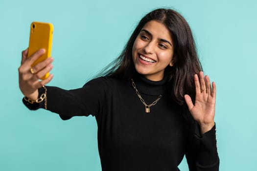 Indian woman traveler blogger, taking selfie on smartphone, communicating video call online with family or friends, live translation stream. Pretty hindu girl isolated on blue background, indoors