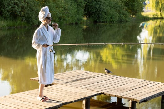Caucasian Woman In White Robe And Towel Is Fishing Holding Spinning Rod On Pier Of Lake Early In The Morning, Green Trees, Nature On Background. Hobby, Leisure Activity, Relaxation. Horizontal