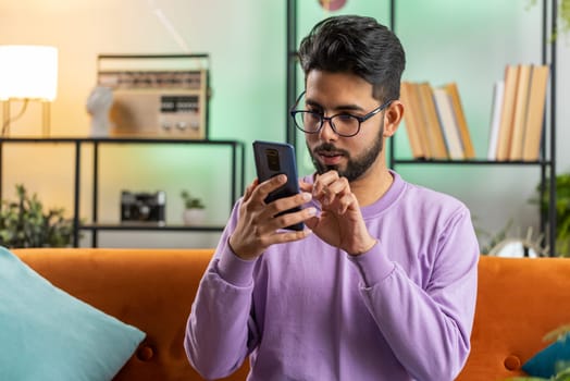 Indian man sitting on sofa uses mobile phone smiles at home living room apartment. Hindu young guy texting share messages content on smartphone social media applications online, watching relax movie
