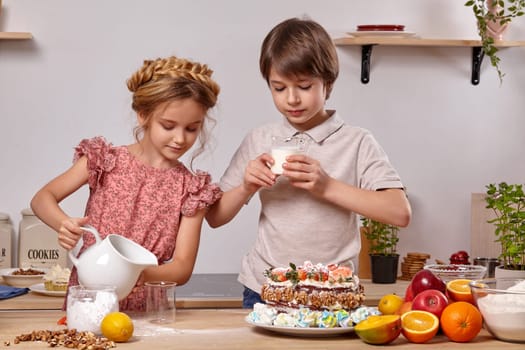 Little brunette boy dressed in a light t-shirt and jeans and a beautiful girl wearing in a pink dress are making a cake at a kitchen, against a white wall with shelves on it. Girl is pouring some milk in a glass and boy is going to drink his milk.