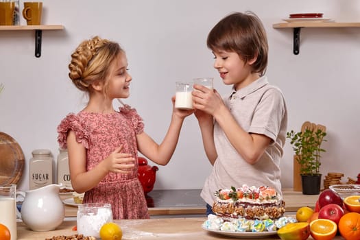 Little brunette child dressed in a light t-shirt and jeans and a beautiful kid wearing in a pink dress are making a cake at a kitchen, against a white wall with shelves on it. Girl and boy cheers with milk.