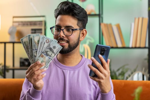 Planning family budget. Smiling indian man counting money cash, use smartphone calculate domestic bills at home room. Joyful hindu guy satisfied of income and saves money for planned vacation, gifts