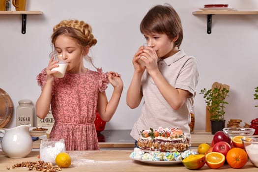 Brunette kid dressed in a light t-shirt and jeans and a pretty girl wearing in a pink dress are making a cake at a kitchen, against a white wall with shelves on it. They are drinking milk.