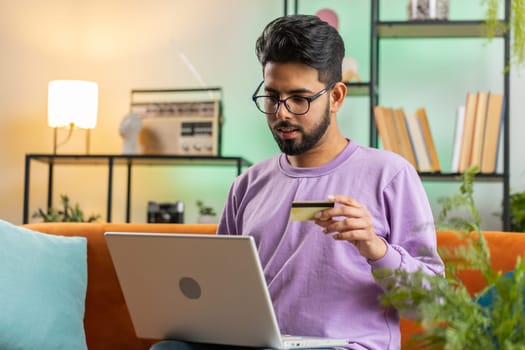 Young indian man using credit bank card and laptop computer while transferring money, purchases online shopping, order food delivery. Portrait of hindu guy at home apartment living room on couch
