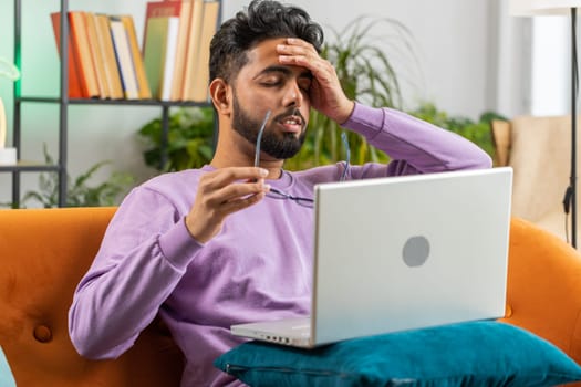Tired freelancer indian man use laptop, suffering from headache problem tension and migraine, stress at home room. Hindu guy works on notebook, sends messages, makes online purchases, watching movies