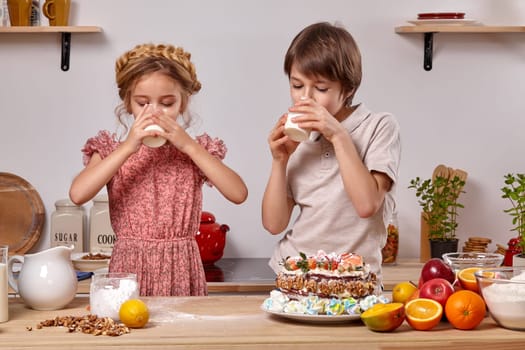 Little pretty boy dressed in a light t-shirt and jeans and a cheerful girl wearing in a pink dress are making a cake at a kitchen, against a white wall with shelves on it. They are drinking milk.